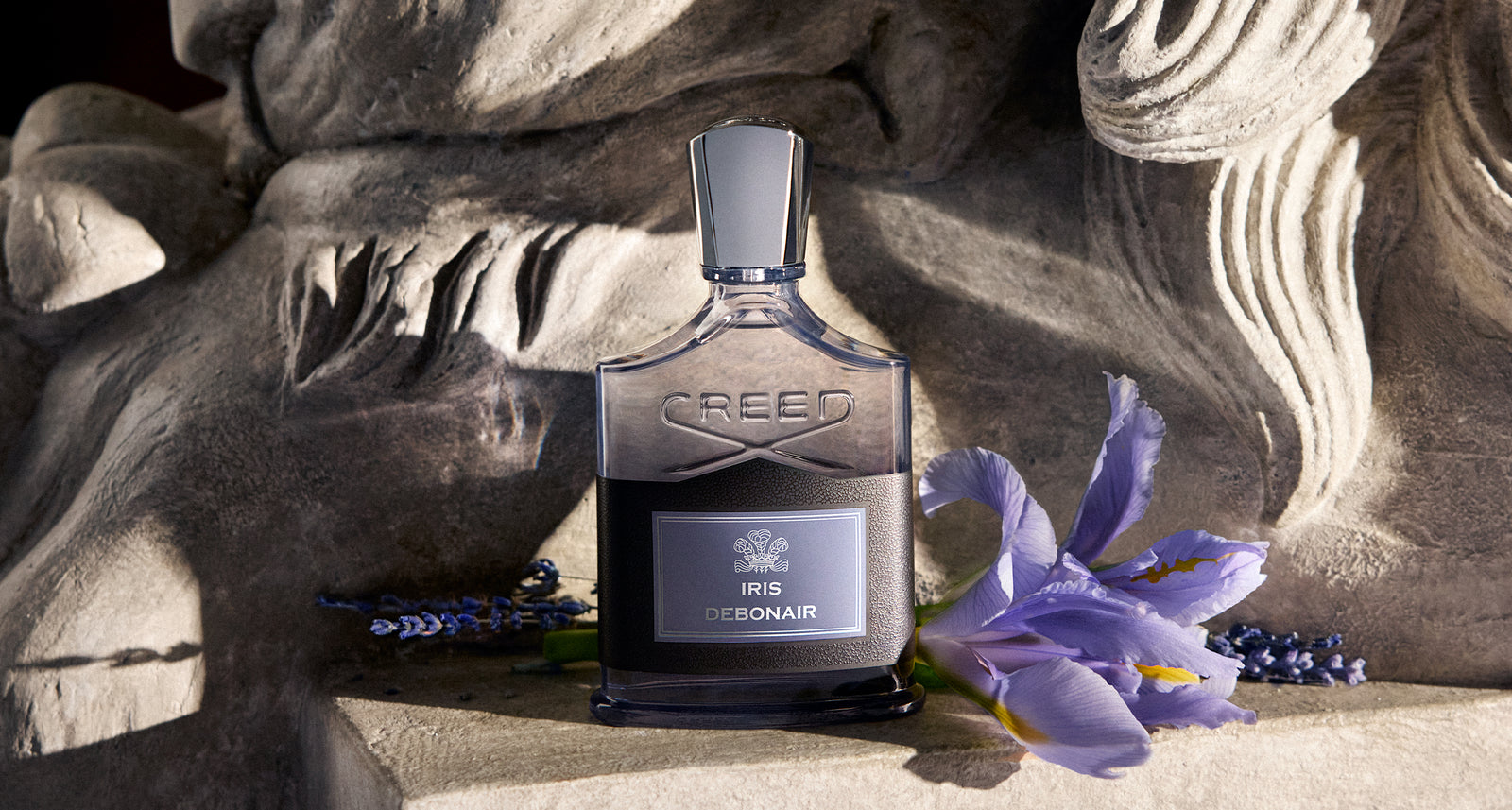 Creed Iris Debonair flacon on a stone plinth beside a purple iris flower